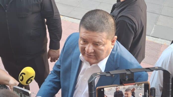 Persona en entrevista con medios en un evento en Chihuahua, vestido con traje azul y camisa blanca, rodeada de reporteros y cámaras, en un entorno urbano.