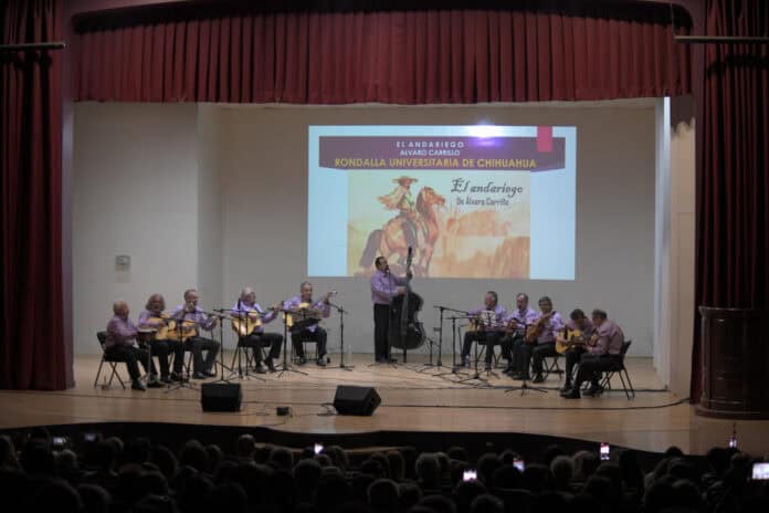 Rondas de músicos en concierto en un auditorio de Chihuahua, presentación de la obra "El Andariego" de Álvaro Carrillo, músicos con instrumentos de cuerda y micrófonos en escenario.