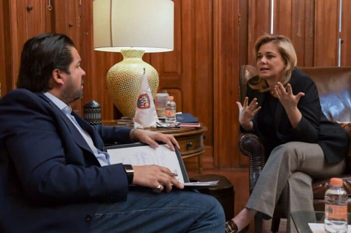 Asesora o reunión entre profesionales en un ambiente formal, en una oficina de madera, con detalles decorativos elegantes y una mesa de trabajo con documentos y agua, en un entorno profesional y de diálogo.