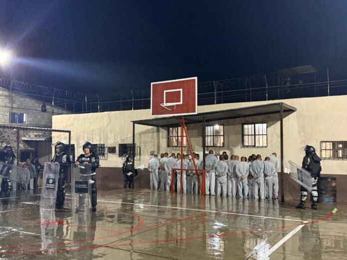 Presos en una cancha de baloncesto, custodiados por policías con escudos y cascos, en un entorno de reclusión con una pared con barrotes y barandales en la parte superior, iluminación nocturna.