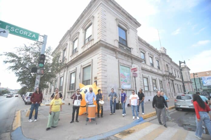 Personas tocando instrumentos en una esquina de la ciudad frente a un edificio histórico, en un evento cultural en Chihuahua, México, promoviendo opciones culturales y comunitarias en la ciudad.