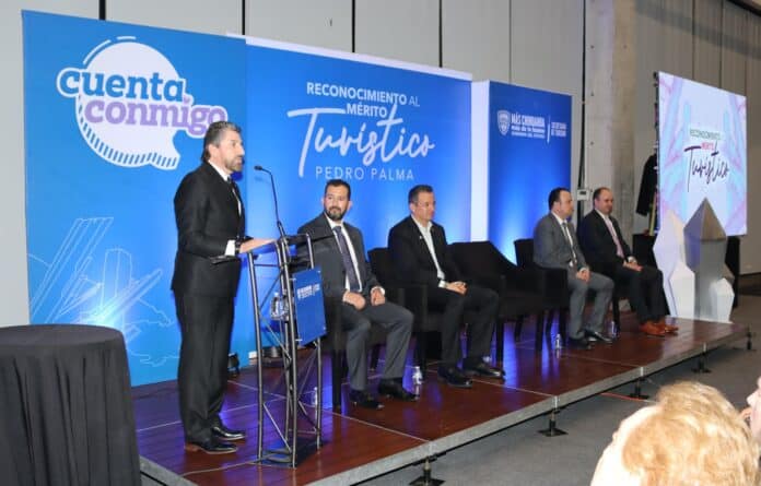 Cita de un funcionario en un evento formal en un escenario con fondo azul y pantallas digitales, resaltando el reconocimiento al mérito turístico en Chihuahua, México.
