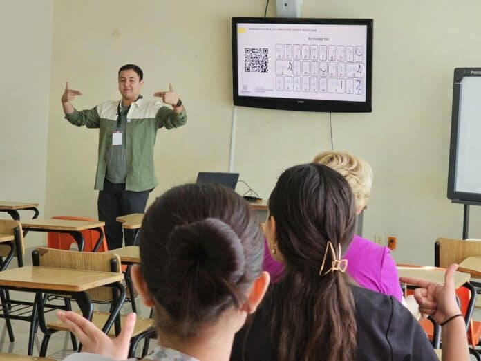 Aprendizaje de lengua de señas mexicana en aula, instructor frente a estudiantes sentados, con pantalla digital mostrando el alfabeto de señas mexicanas, en un entorno educativo inclusivo.