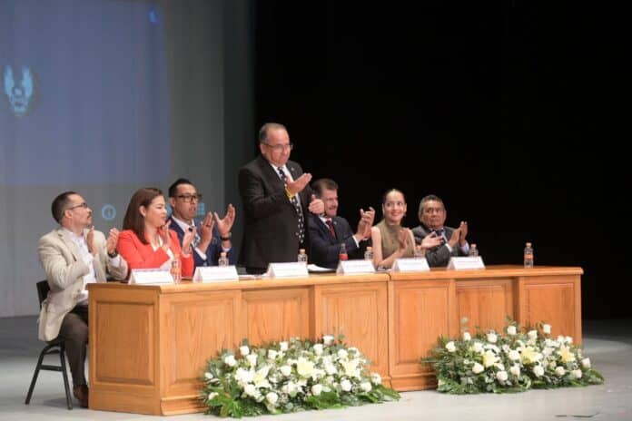 Profesionales y autoridades en una conferencia o panel de discusión, en un evento formal con decoraciones florales y fondo de pantalla institucional, promoviendo liderazgo y participación en la comunidad.
