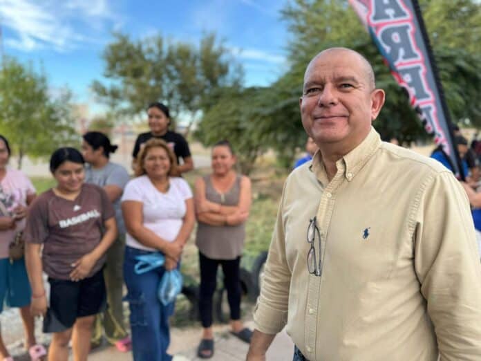 Hombre sonriendo en evento al aire libre con grupo de mujeres en fondo, en un parque con árboles y cielo despejado.