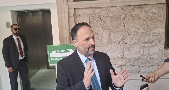 Candidato en una reunión oficial, hablando con medios de comunicación en un entorno urbano, con fondo de muro de piedra y señal de ruta de evacuación, promoviendo temas de interés público y eventos políticos.