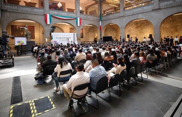 Amplio auditorio lleno de asistentes en un evento institucional o académico, con una presentación en pantalla gigante y decoraciones patrióticas, en un espacio con arquitectura de estilo colonial mexicano.