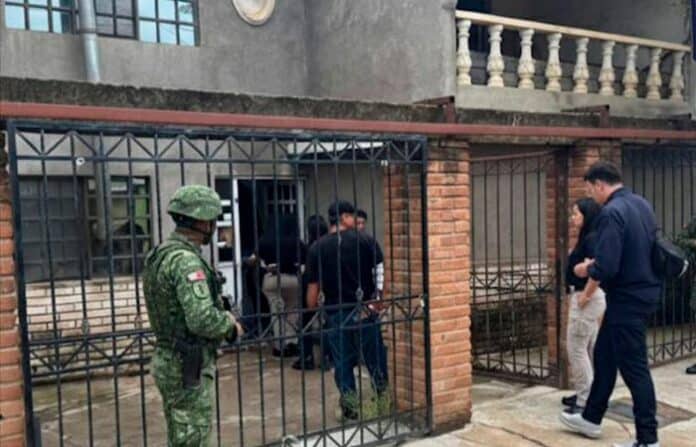 cateocasaparral Soldado en uniforme en una reja de entrada a una casa en Chihuahua, México. Personas conversando en la calle frente a la vivienda, en un entorno urbano, relacionado con seguridad y protección en la región.