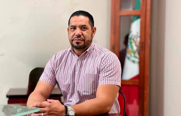parralpriapo Profesional mexicano sentado en oficina, uso de camisa de rayas y reloj, con bandera de México en el fondo, promoviendo confianza y liderazgo para sitios como soychihuahua.mx.