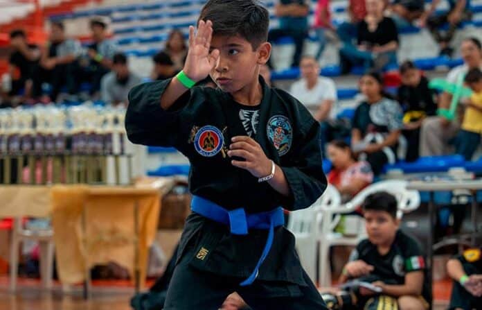 Niño practicando karate en competencia deportiva en Chihuahua, en un torneo de artes marciales con público y otros niños en el fondo.
