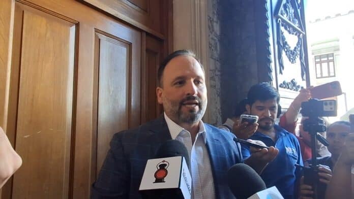 Abogado o funcionario hablando en una rueda de prensa con medios de comunicación presentes, en un entorno con detalles de madera y decoración tradicional mexicana, en Chihuahua.