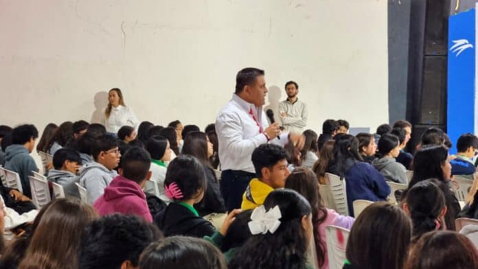 Alt: Hombre con micrófono dirigiéndose a un auditorio escolar lleno de estudiantes en un evento académico en Chihuahua.