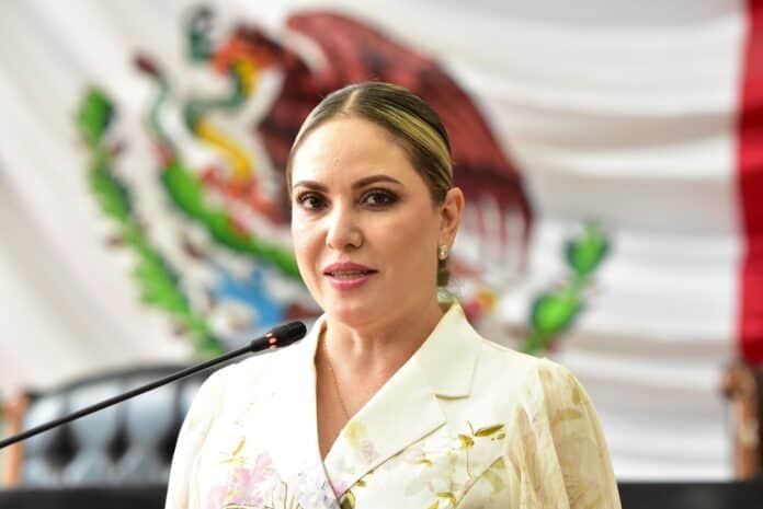3a21023c-7fcf-4f54-848f-436231e20080 Comunicadora gubernamental en evento oficial con bandera de México de fondo.