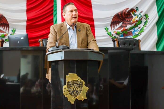 Diputado en sesión del Congreso de Chihuahua con bandera de México y escudo estatal de fondo.