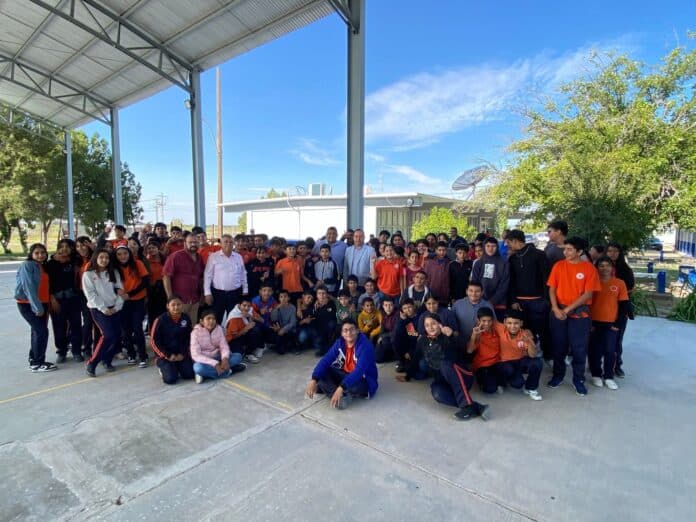 Grupo de estudiantes y docentes en evento escolar en Chihuahua, México, bajo un domo de metal y con un cielo despejado, promoviendo educación y desarrollo juvenil en la región.