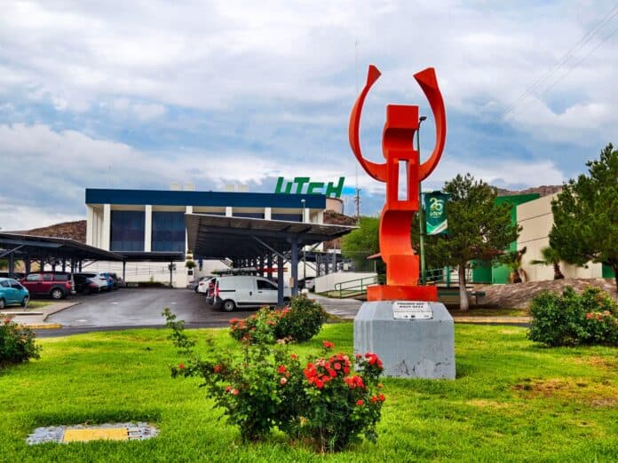 Estatua moderna de color naranja en el campus de UTECh, rodeada de áreas verdes y flores, con estacionamiento y edificio en el fondo bajo cielo nublado, representando innovación y educación en Chihuahua.
