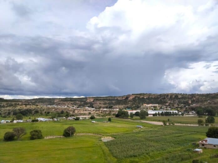 Nube oscura sobre la campiña de Chihuahua, con campos verdes, casas y cerros en el fondo, mostrando la belleza natural y rural del estado.