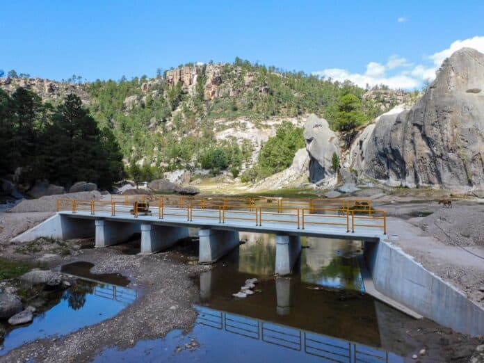 IMG-20251007-WA0062 Puente peatonal en un río del paisaje natural de Chihuahua, rodeado de formaciones rocosas y vegetación, ideal para turismo y conexión en áreas naturales.