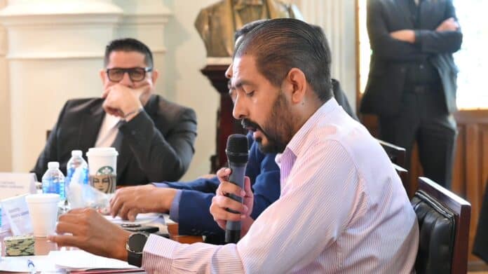 Audiencia en una sala formal con hombre hablando con micrófono, otros asistentes en la mesa, ambiente profesional.