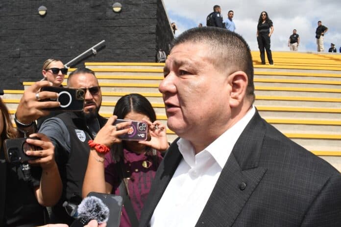 IMG-20251009-WA0071 Archivo de imagen de un hombre en traje oscuro siendo entrevistado por periodistas, con varios reporteros tomando fotos y videos en el fondo, en un entorno de escalones y cielo nublado.