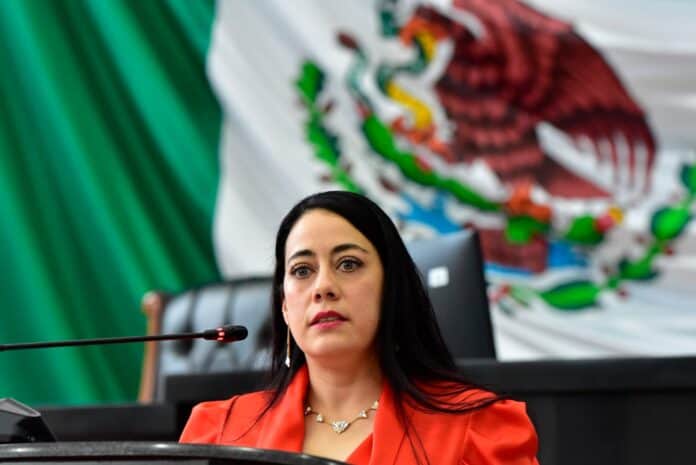 México, mujer política, en sesión legislativa con bandera de México de fondo.
