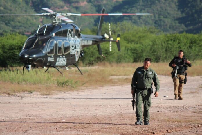 IMG-20251011-WA0001 Helicóptero de la policía de Chihuahua en entrenamiento o patrulla en áreas rurales. Policía estatal en operación con equipo táctico y armas, en una escena en terreno abierto con vegetación y montañas de fondo.
