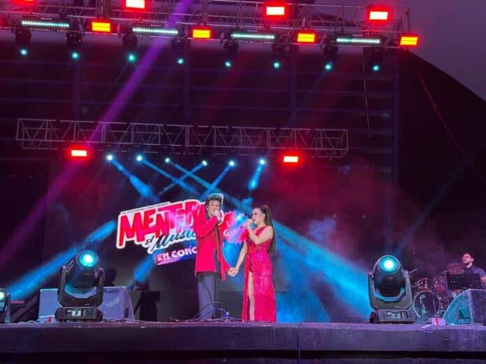 Pareja cantante en escenario durante concierto en Chihuahua con iluminación colorida y máquina de luces, promoviendo entretenimiento musical en eventos culturales del estado.