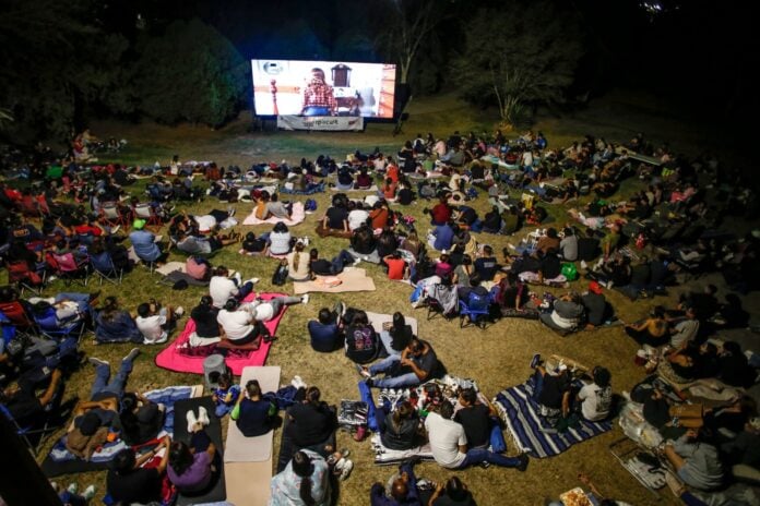 Disfruta de una noche de cine al aire libre en Chihuahua, con mucha gente viendo una película en una pantalla gigante, sentados en mantas y sillas, en un ambiente familiar y cultural.
