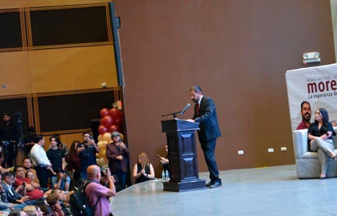 Padrino dando discurso en un escenario durante un evento formal con audiencia atenta y varios reporteros captando el momento.