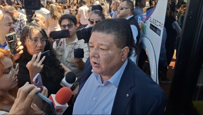 Imagen de un político, rodeado de periodistas y reporteros, en un evento público en Chihuahua, México, con muchas personas en el fondo.
