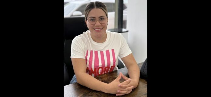 Chica sonriendo en una cafetería, usando gafas y camiseta blanca con letras rosas, en un ambiente urbano y moderno en Chihuahua, promoviendo estilo de vida saludable y bienestar.