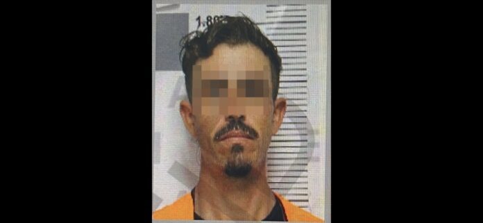 Screenshot_20251003_113904_WhatsApp Hombre en retrato policial con bigote y barba delgada, usando uniforme naranja, en fondo de línea de medición, en un contexto que puede estar relacionado con temas de seguridad o justicia en Chihuahua.