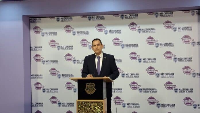 Armado con traje y corbata, un funcionario del gobierno del estado de Chihuahua pronuncia un discurso frente a un fondo con logotipos oficiales de "Más Chihuahua" y "Cuenta Conmigo".
