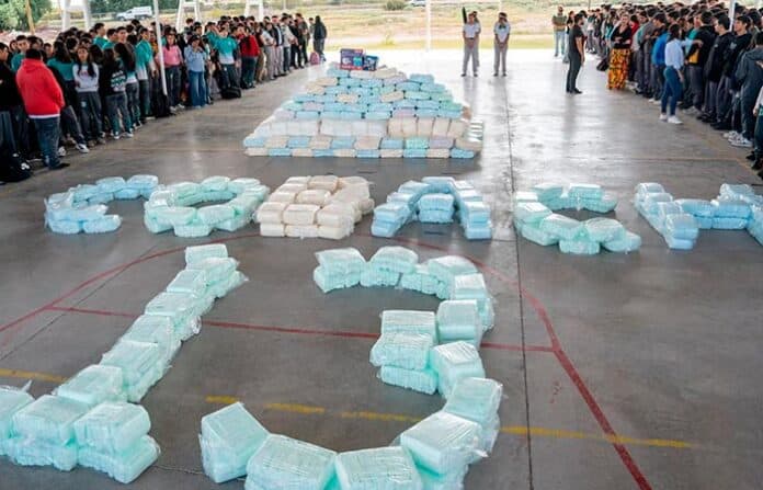 difcobachdeli Blocs de drogas formando la palabra "PAZ" en una cancha escolar, con personas observando en el fondo, en Chihuahua, México. Campaña contra el narcotráfico y promoción de la paz en la comunidad.