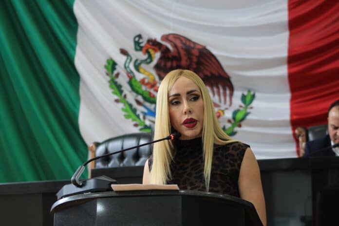 Imagen de una mujer en un acto oficial en una sala con la bandera de México de fondo, destacando la participación política y el compromiso con el país.