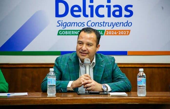 memofogon Bajo el sol, un hombre en una conferencia política en Chihuahua, con carteles del gobierno local y agua en la mesa, promoviendo el desarrollo y la gestión gubernamental en la región.